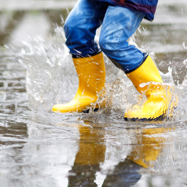 Close-up of kid wearing yellow rain boots and walking during sleet, rain and snow on cold day. Child in colorful fashion casual clothes jumping in a puddle. Having fun outdoors.